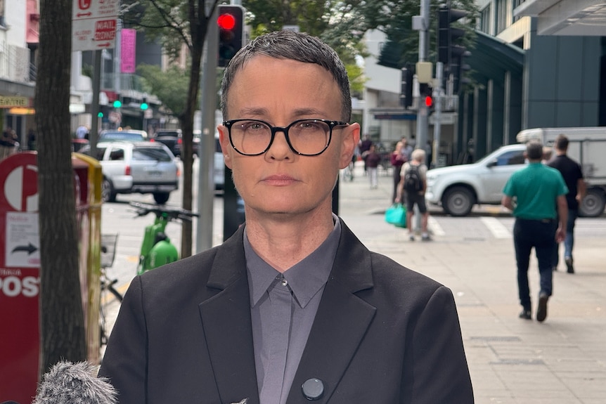 A woman with short hair and glasses in a dark suit standing on a city street