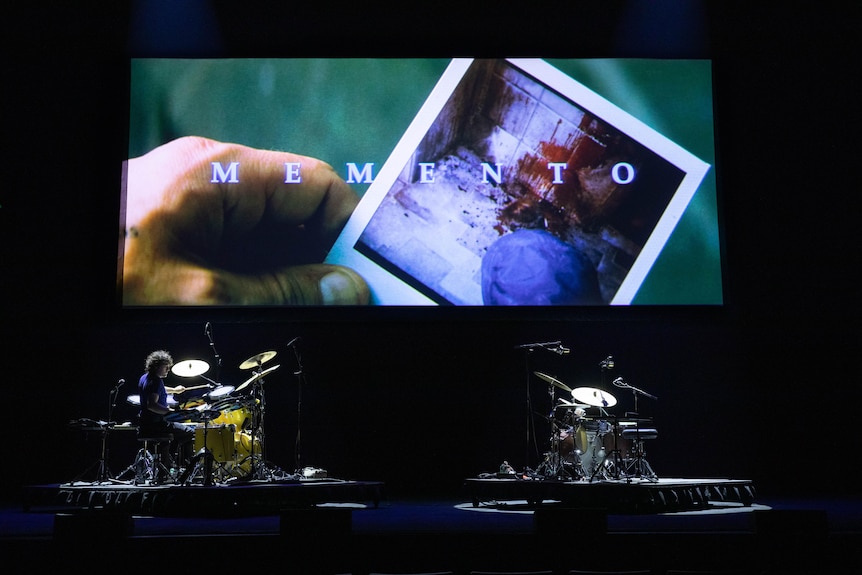 Cinema screen shows title sequence of 2000 film Memento. Onstage are two large drumkits, one played by curly-haired man