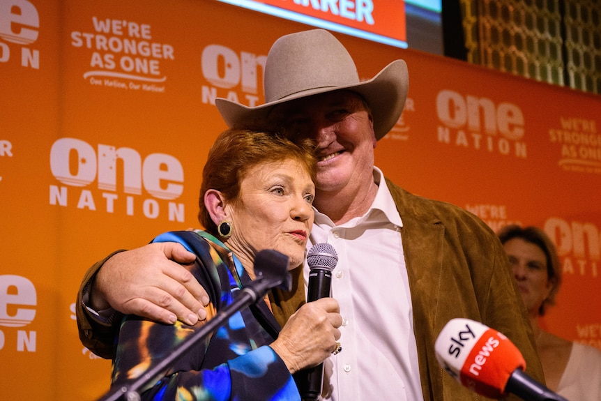 A man in an Akubara wraps his arms around a woman with a microphone.