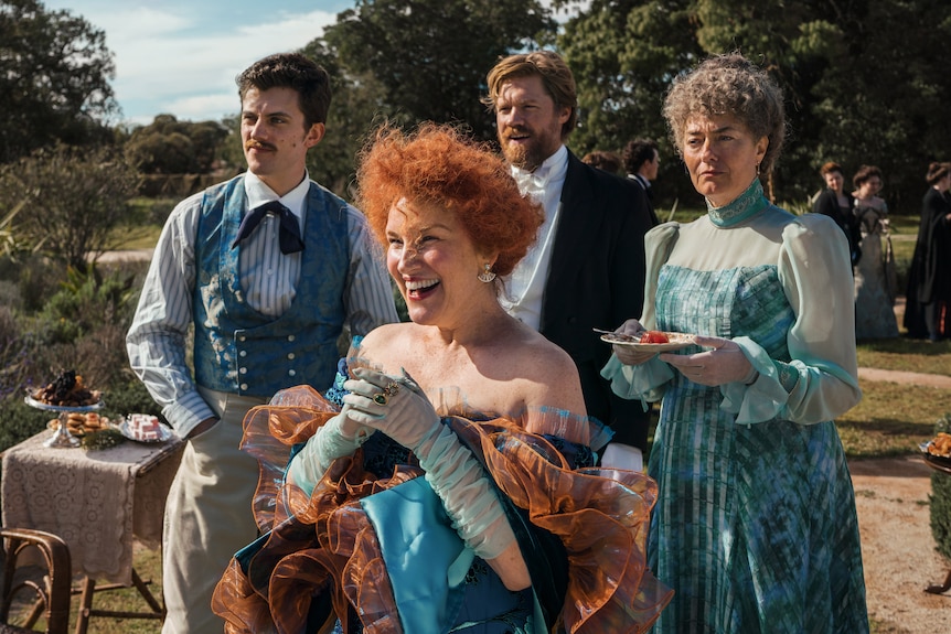 A woman with curly red hair and period clothing and gloves laughs, two men and a woman behind her