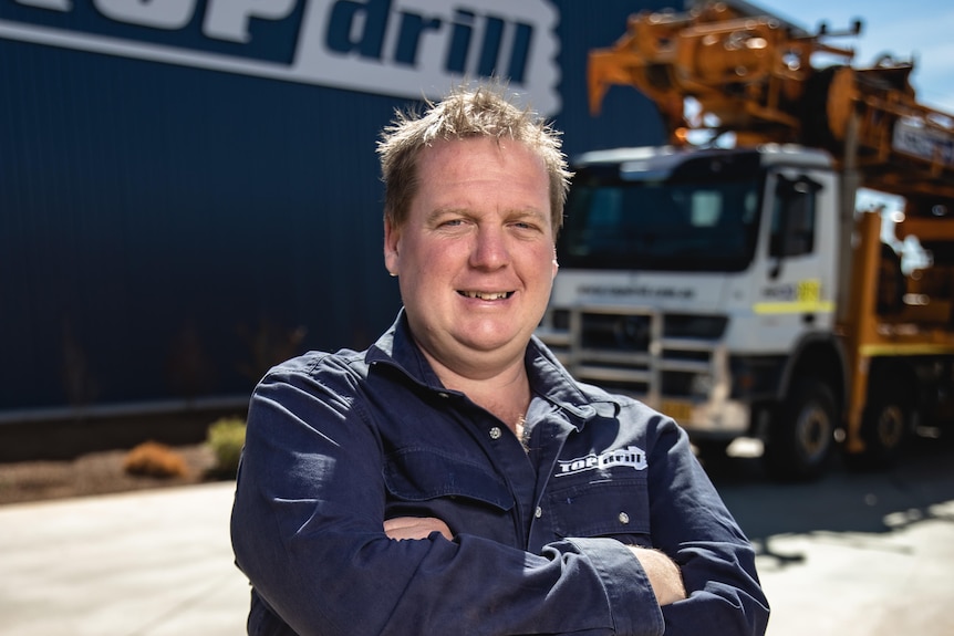 A smiling, middle-aged man in a dark work shirt stands near a truck and a warehouse on an industrial site.