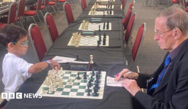 A young boy wearing a white shirt and colourful glasses, with short dark hair sits on a red chair at a table opposite a man with grey hair, wearing a suit and glasses, with a newspaper rolled up in his pocket. They are playing chess. There are many other chess boards and chairs lined up along tables in the room.