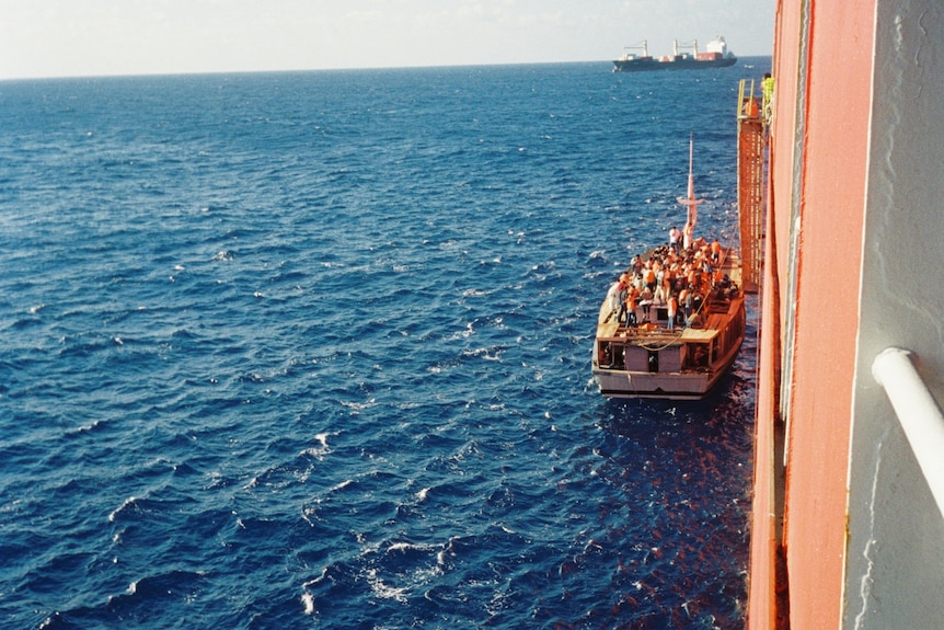 A small wooden boat pulls up to the side of a large freight ship.