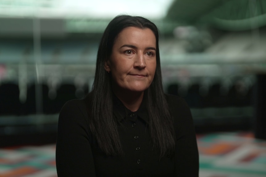 A woman with long dark hair wearing a black shirt sits in a room overlooking a large football stadium.