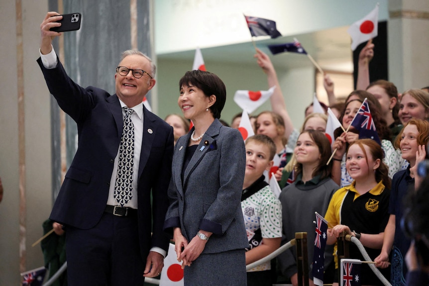 Albanese and Takaichi take a smiling selfie photo in front of a crowd of children waving Japanese and Australian flags.