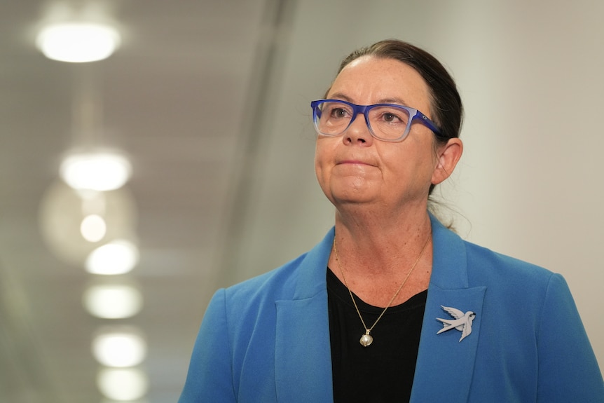 A dark-haired woman in glasses wears a blazer with a brooch as she stands in a corridor.