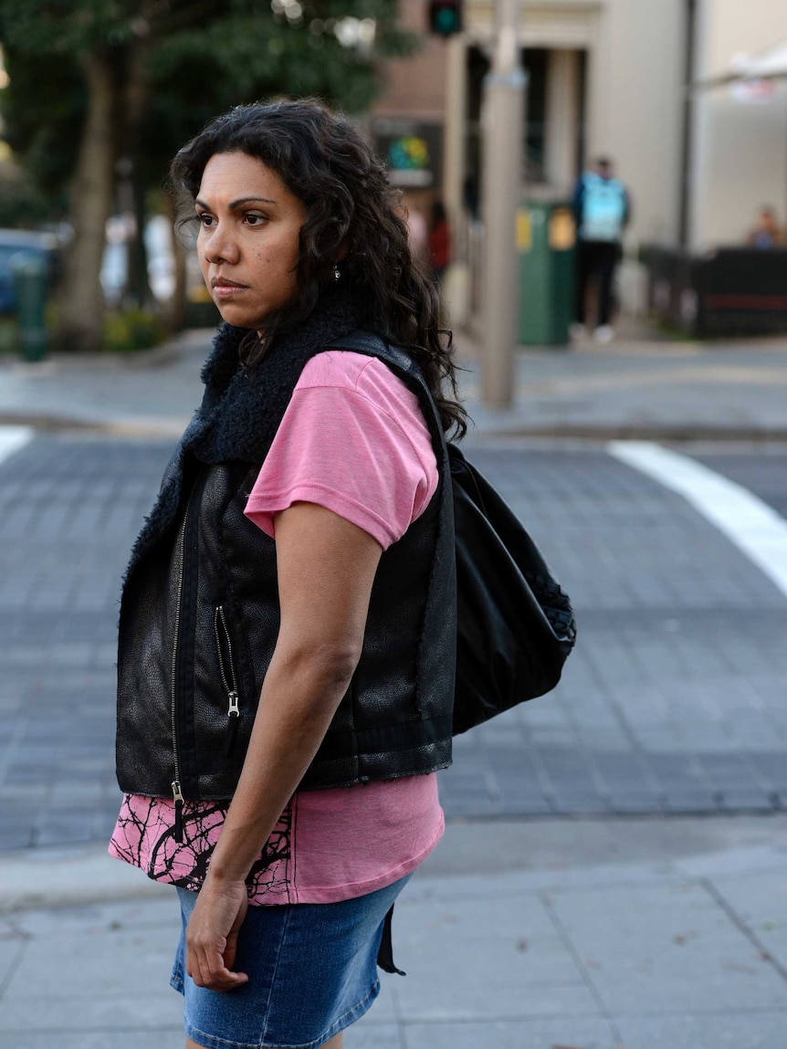 A woman with long, dark hair wears a brightly coloured shirt and dark vest while standing on a city street.
