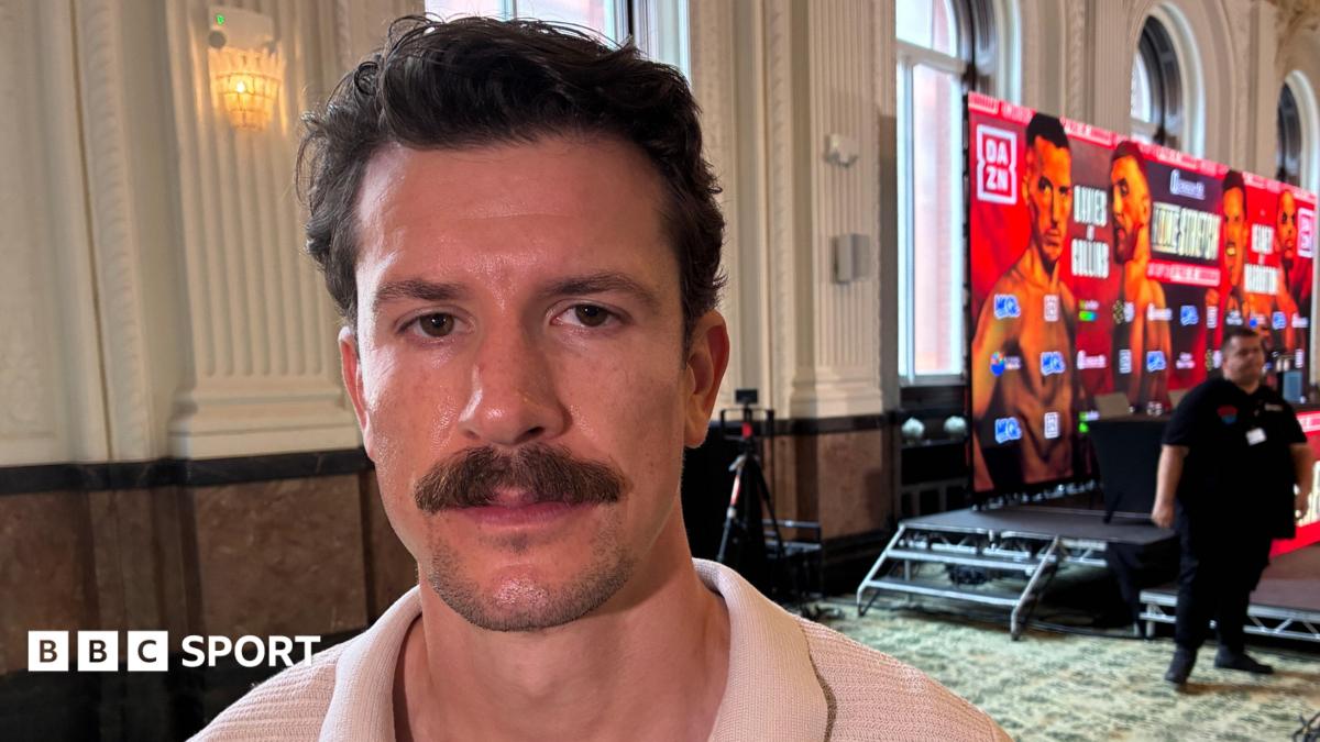 Boxer Nathan Heaney, who has dark hair and a moustache, stands to the side of a promotional dais advertising his next bout. A security man stands in the background to his left
