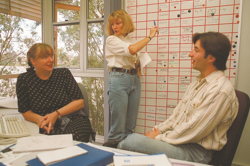 A 1990s office with a woman standing at a white board holding a pen. She looks over at a man and woman who are seated.