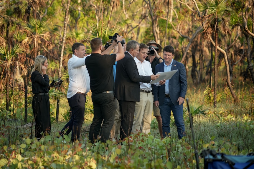 A group of three men in business wear stand in tropical savanna bushland, looking at paperwork surrounded by a press pack.