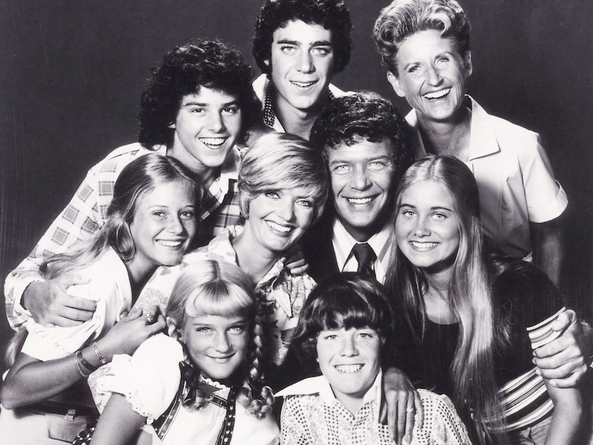 A black and white photo of a family of eight with their housekeeper, mix of blonde and brown hair, smiling, cuddled together.