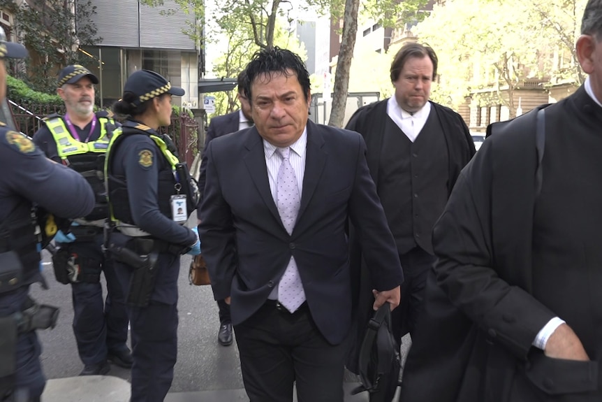 A dark haired man in a dark suit, white shirt and light purple tie walks past police beside barristers.