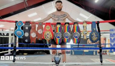 Jason Cunningham is wearing dark shorts and white boots. He has short brown hair, a brown beard and tattoos across his chest. He stands inside a boxing ring behind a row of championship belts draped across the top rope. A large silver trophy is positioned on the ring floor in front.