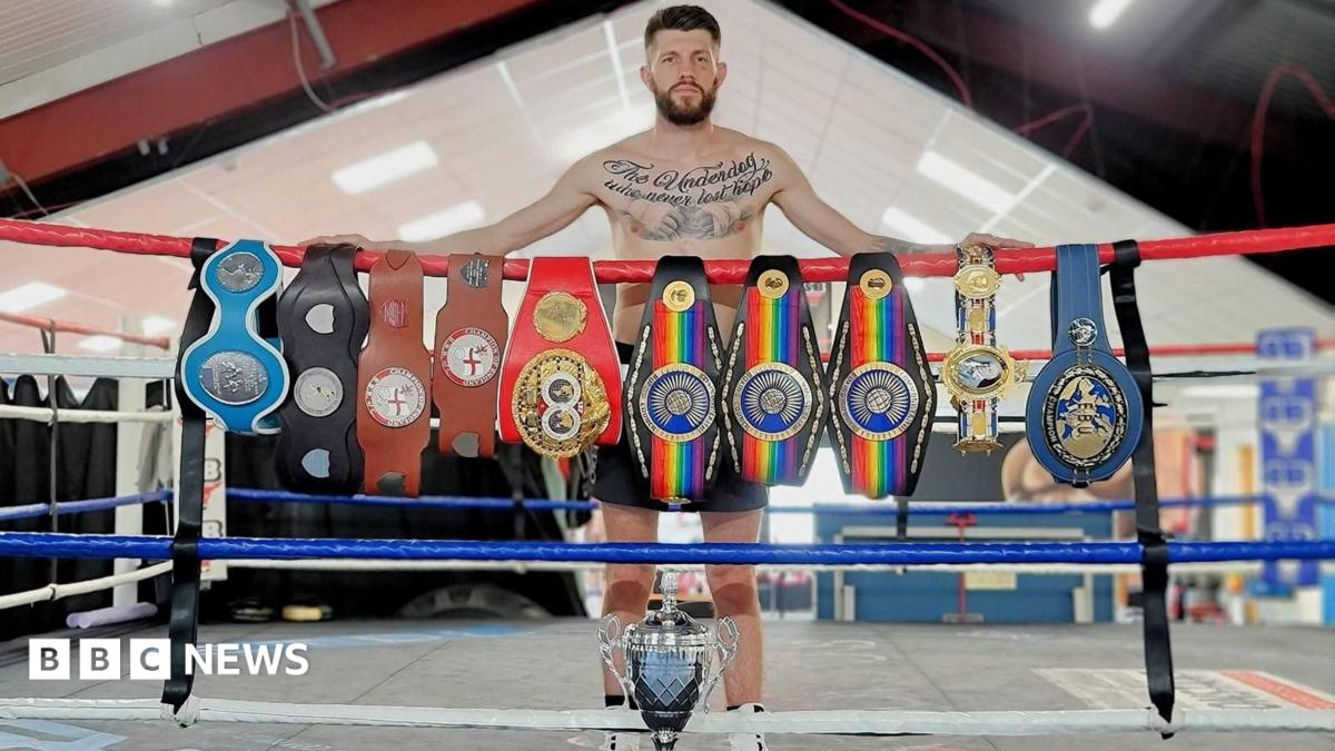 Jason Cunningham is wearing dark shorts and white boots. He has short brown hair, a brown beard and tattoos across his chest. He stands inside a boxing ring behind a row of championship belts draped across the top rope. A large silver trophy is positioned on the ring floor in front.
