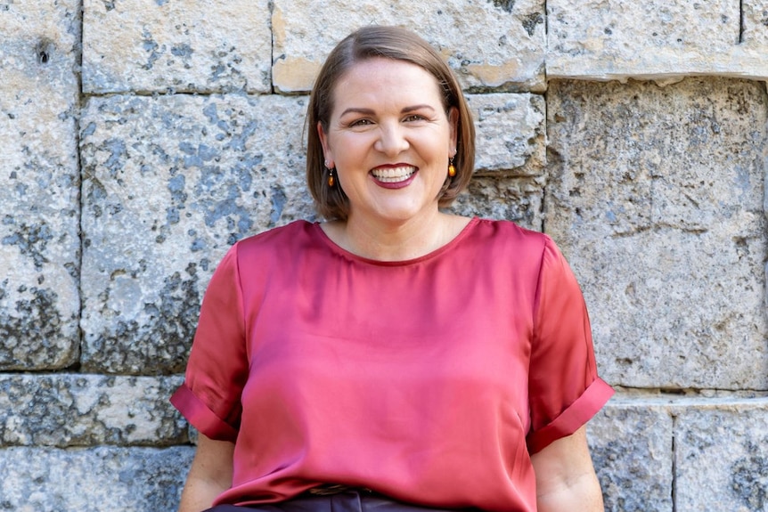 A woman wearing a pink shirt smiles against a brick wall
