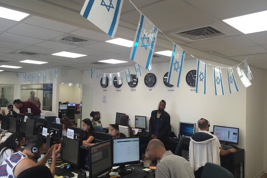 People with blurred faces work at a call centre with small Israeli flags hanging from the roof