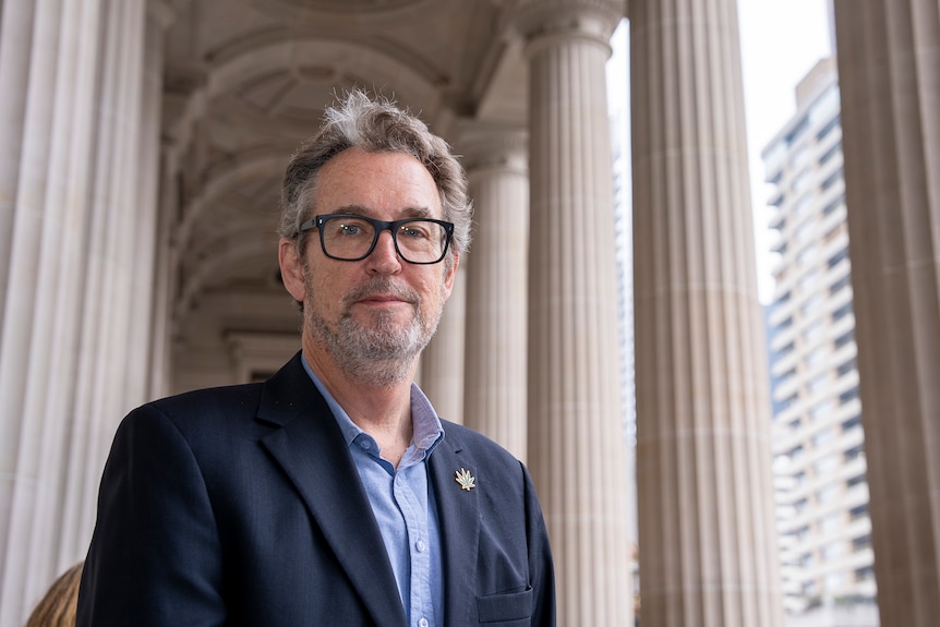 A man wearing glasses and a blue blazer stands near sandstone columns of a building.