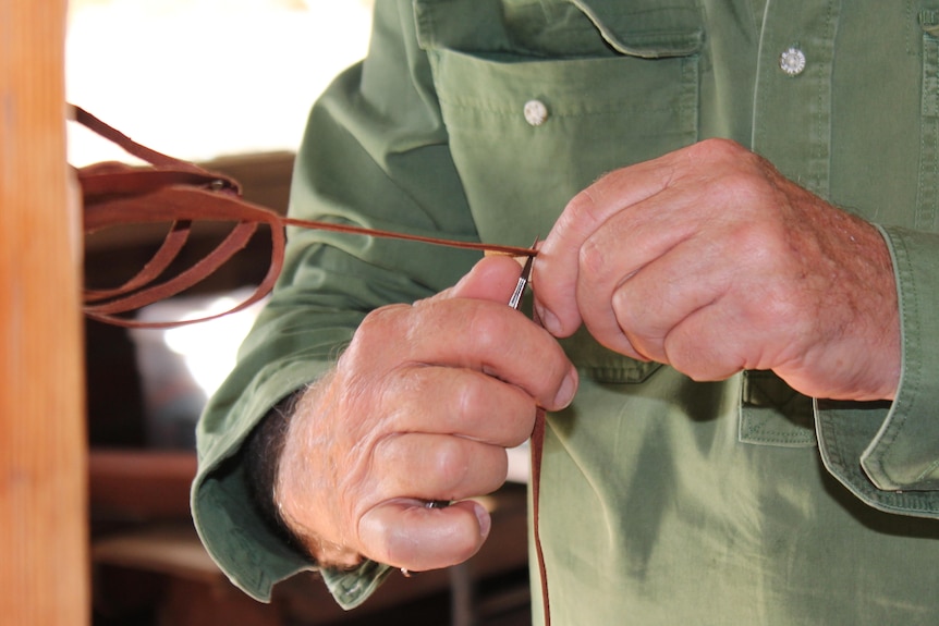 An older pair of mens hands holding a thin strand of brown leather and a sharp silver knife.
