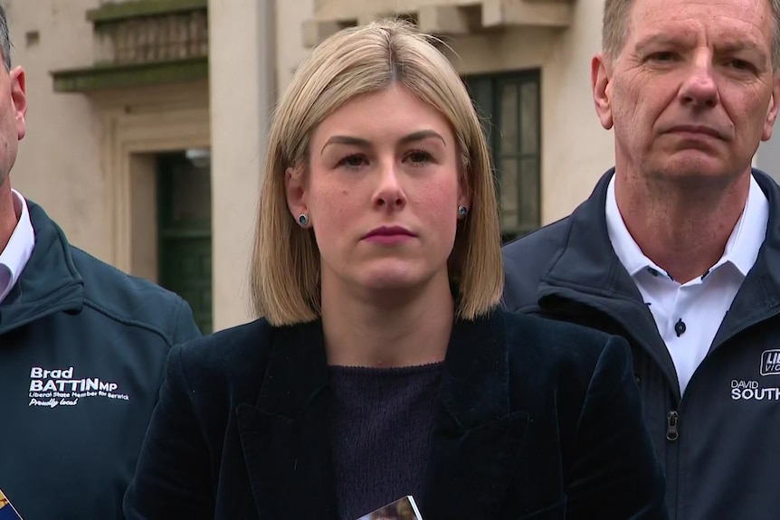 A woman with chin length blonde hair in a black jacket stands in front of two men in blue jumpers.