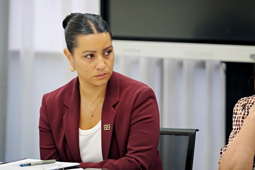 A woman with her hair parted in the middle and tied back, wearing a maroon blazer, looks off to the right