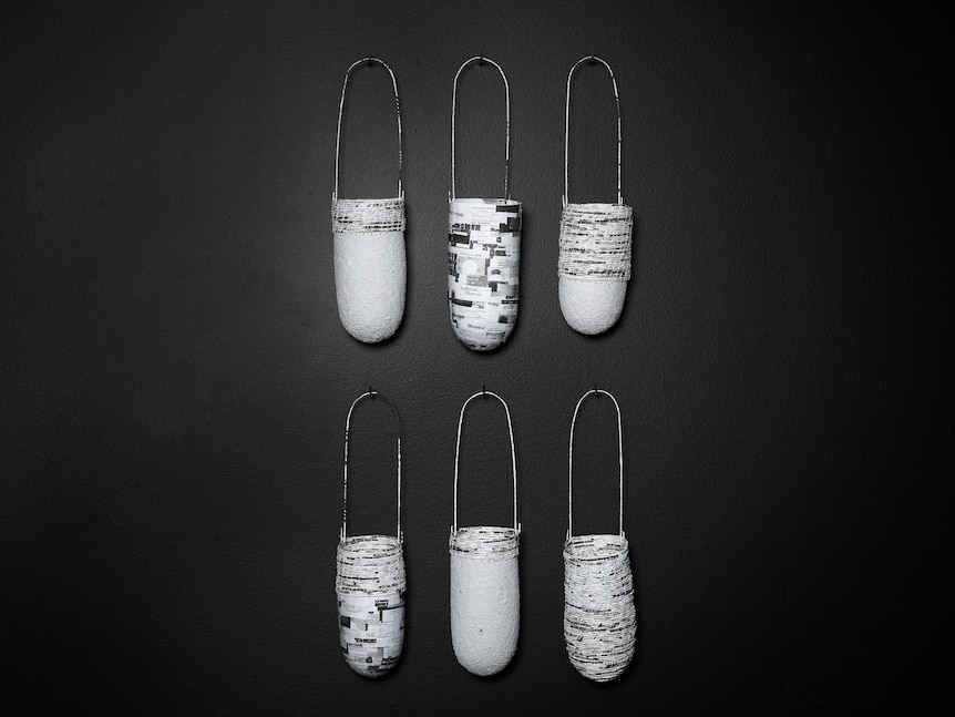 Six woven bags hang from a wall. They're white and black and made from strips of paper turned into string.