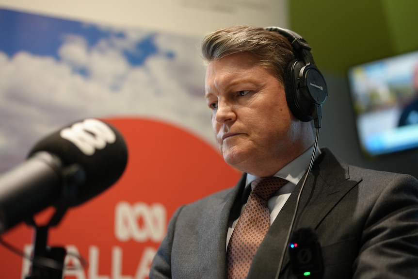 A man in a grey suit sits in front of a microphone with an ABC logo on it with headphones on.