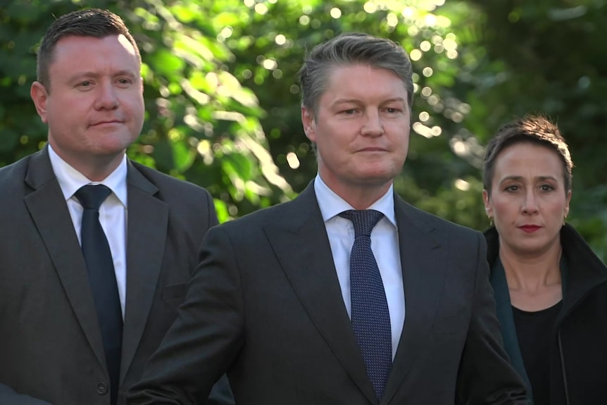 Two men in grey suits, white shirts and blue ties and a woman with short dark hair dressed in black standing near bushes.