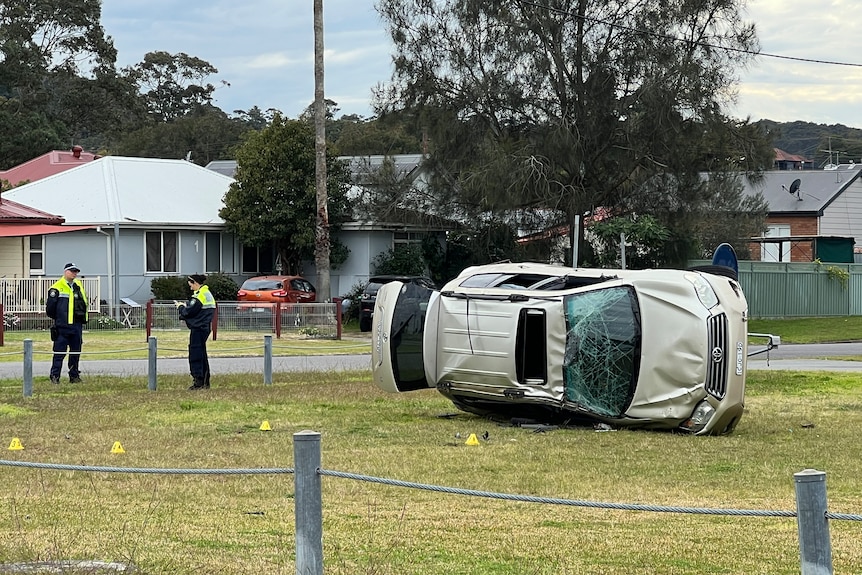 Two police officers stand next to a field. A rolled car with a smashed windscreen lies in the middle.