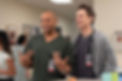 Two smiling doctors in a hospital hallway, one in green scrubs gesturing and one in a gray cardigan with a stethoscope.