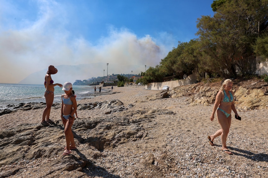Tourists on a beach with a large smoke plume seen in the far distance.
