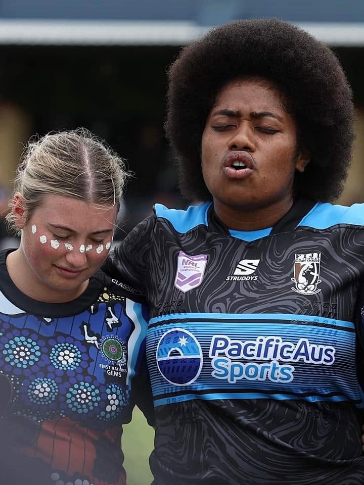 A Fijian woman with a traditional Fijian hairstyle has her eyes closed with her arms around another girl as she sings or prays
