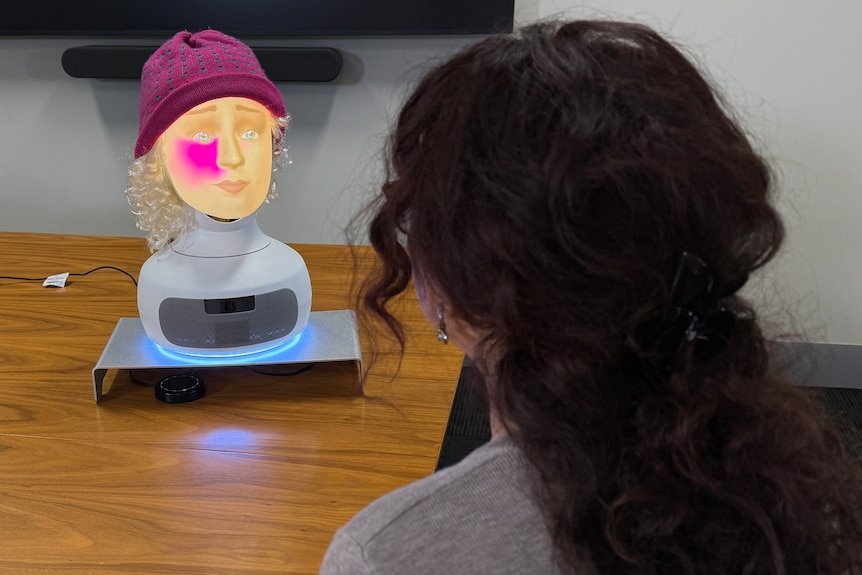 A robot with an illuminated human-like face sits on a table, with a pink glow on its cheek and wearing a pink hat. Woman's head 