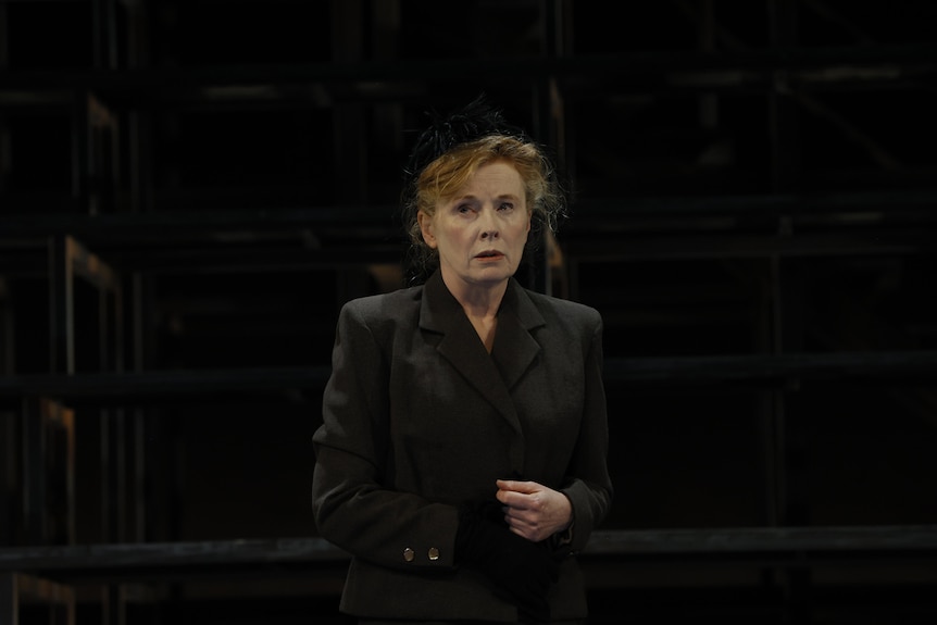 On stage, Alison Whyte, mid-50s, red hair tied back, dressed in a black hat and blazer, looks troubled.