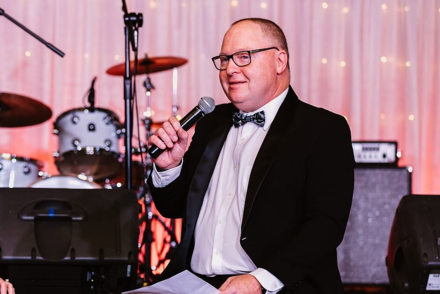 A man in black tie speaks into a microphone while sitting on a stage near a drum kit.