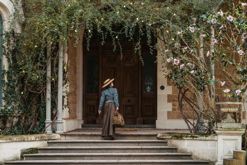 A woman dressed in 1900s clothing stands at the grand entrance of a mansion.