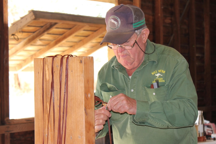 A older man in a cap and glasses works with long strips of leather.