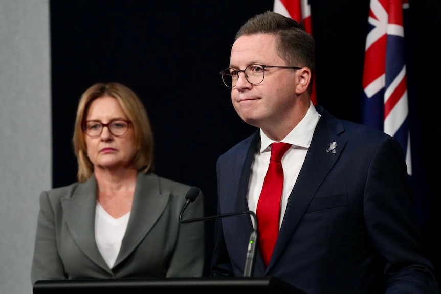 Jacinta Allan and Anthony Carbines stand together in a room in front of Australian flags.