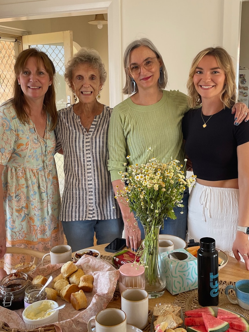 Four woman stand in a line smiling. In front are flowers and a tea set