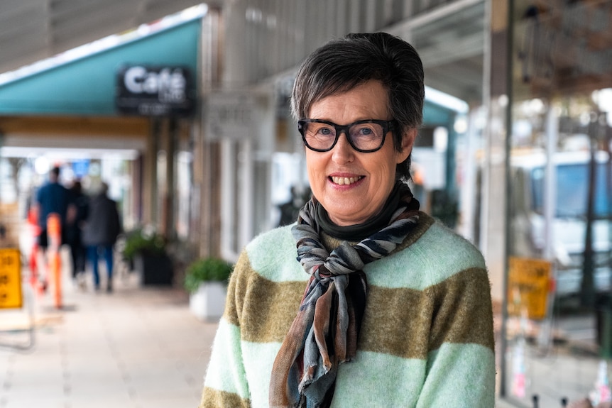 A lady wearing a green jumper smiles from the footpath of a regional town in front of her small business.