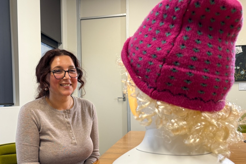 A woman wearing glasses looks toward a doll-like head that can be seen from behind. It has blonde curly hair, wearing a pink hat