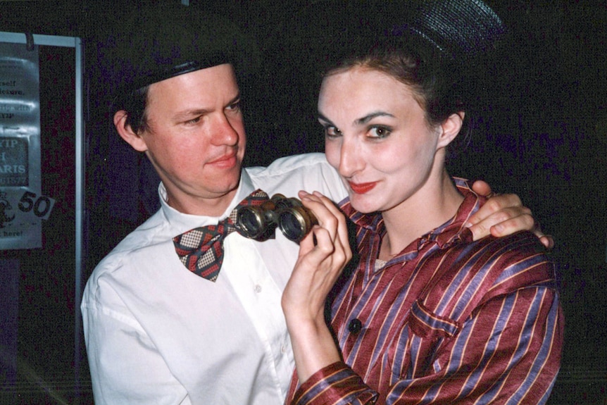 A man and woman in theatre costumes including hats. Woman holds up small binoculars