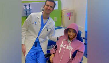 A male doctor in a white lab coat smiles with a young patient in a pink hoodie in a wheelchair in an exam room.
