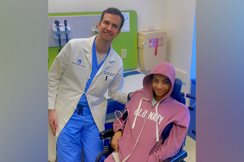 A male doctor in a white lab coat smiles with a young patient in a pink hoodie in a wheelchair in an exam room.