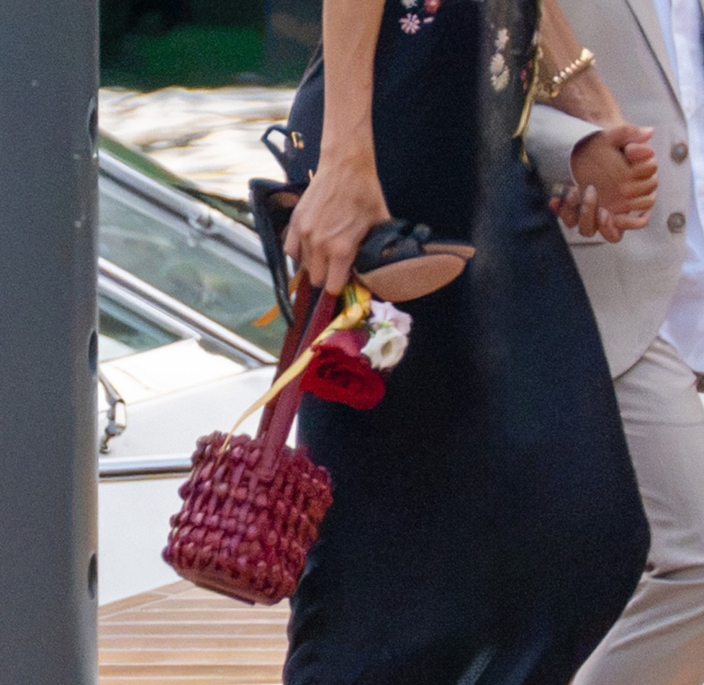 A woman in a black dress holds a woven red bag with flowers, walking next to a man in a tan suit, with a boat in the background.