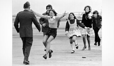 A young girl runs with open arms toward a uniformed man, likely her father, at an airport while other smiling family members follow joyfully behind her.
