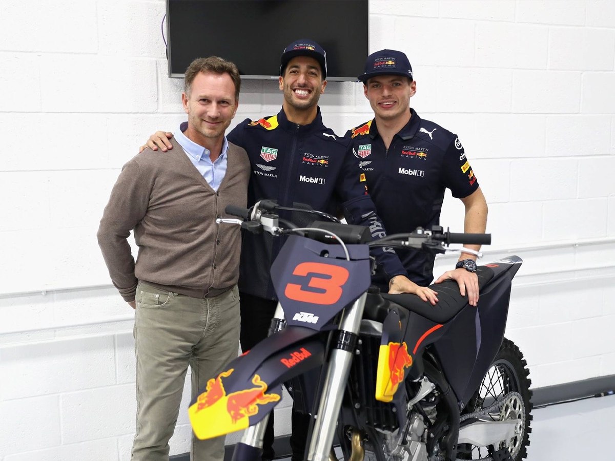 Daniel Ricciardo and two others pose with a Red Bull KTM motorcycle indoors, all smiling.