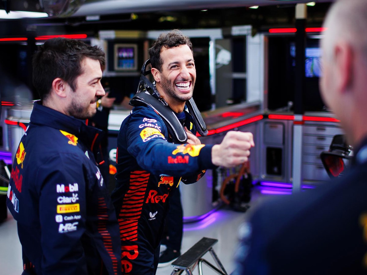 Daniel Ricciardo smiling in Red Bull Racing suit, surrounded by team members in a brightly lit garage.