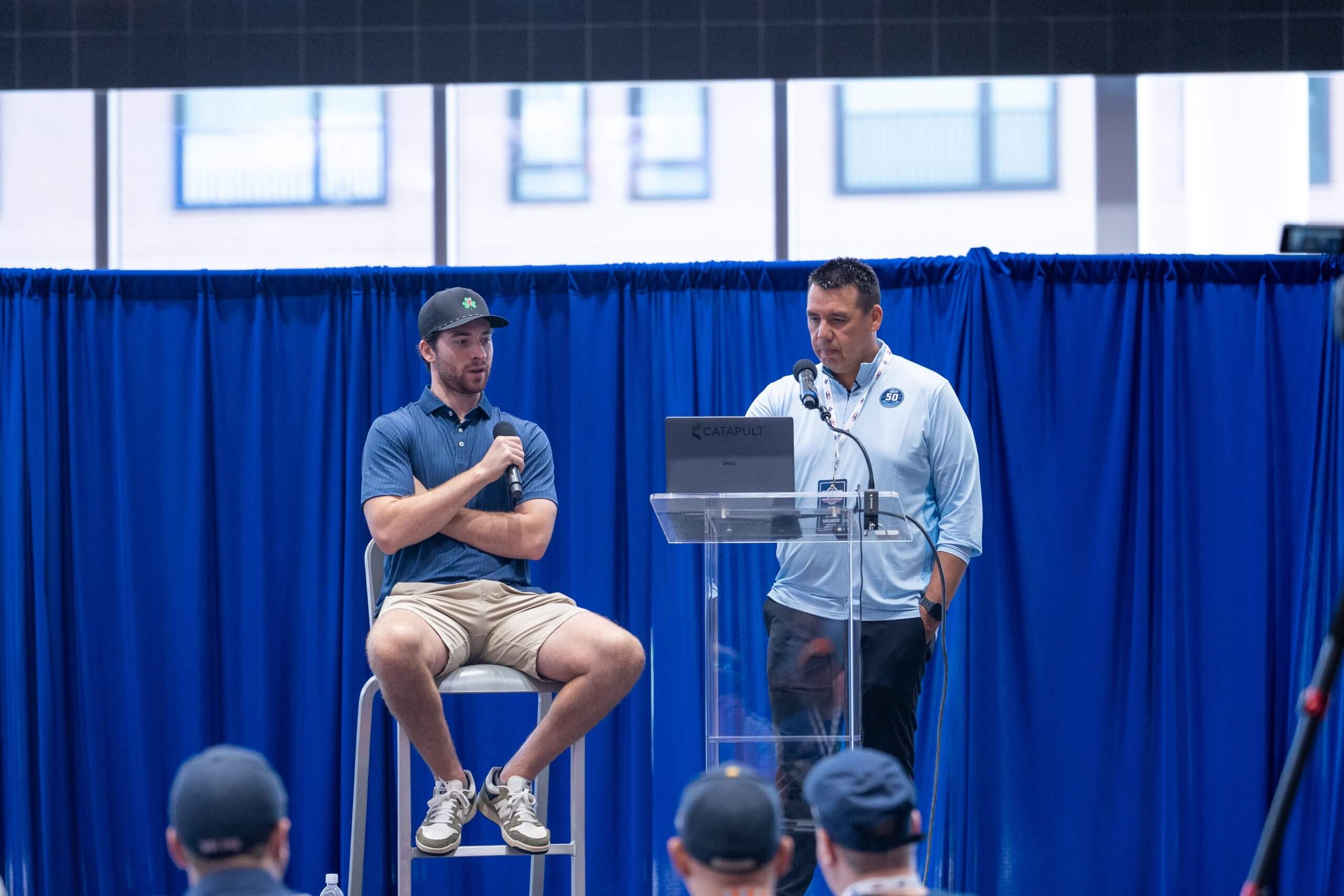 On a stage in front of a blue background, Bruins goalie Jeremy Swayman sits on a chair with a microphone next to Maine goalie coach Alfie Michaud, who is looking at a laptop.