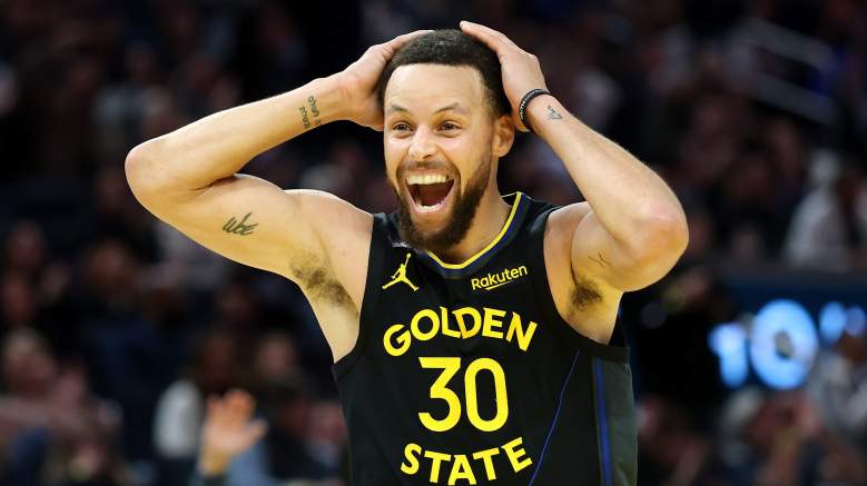 Stephen Curry smiles with both hands on his head while wearing a black Golden State Warriors uniform.