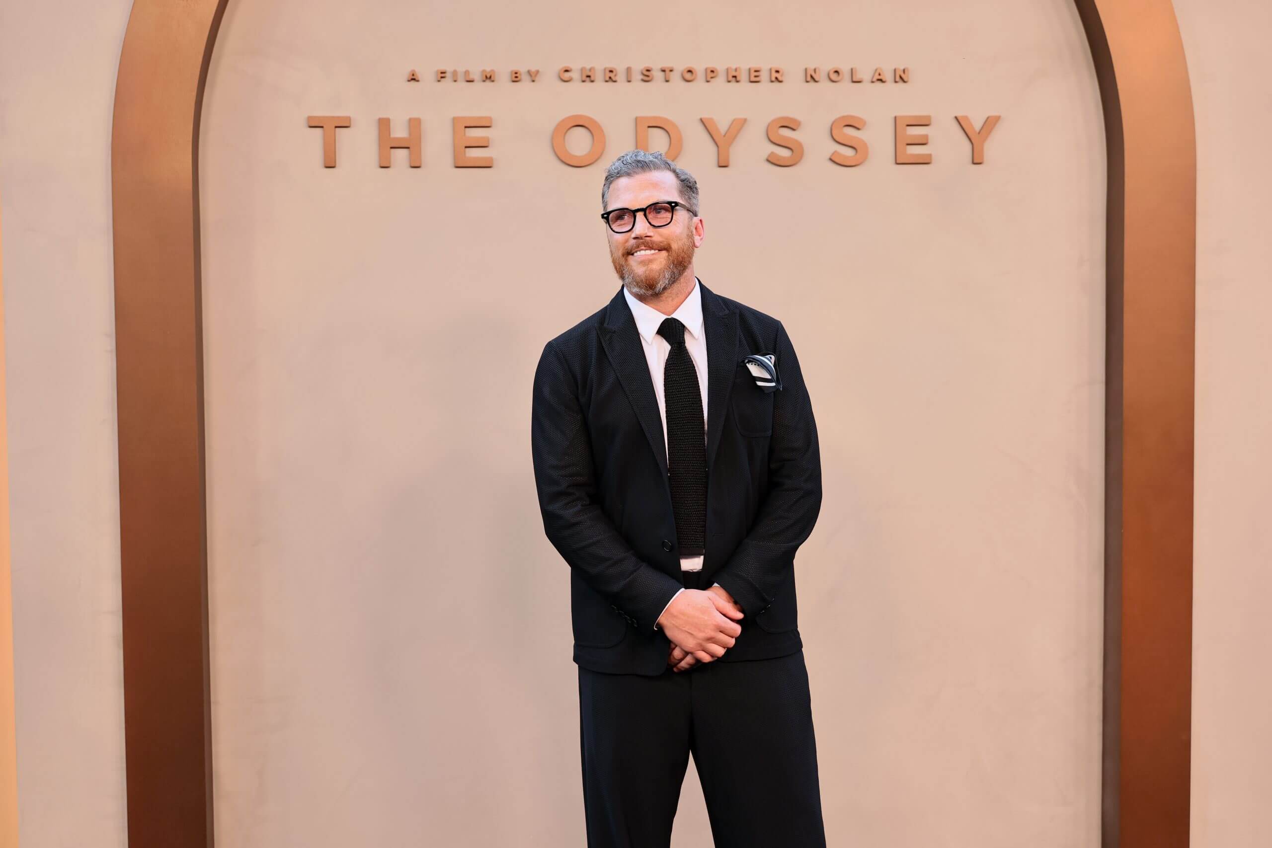 Sean Avery smiles in a suit with hands clasped in front of him, in front of a "The Odyssey" backdrop.